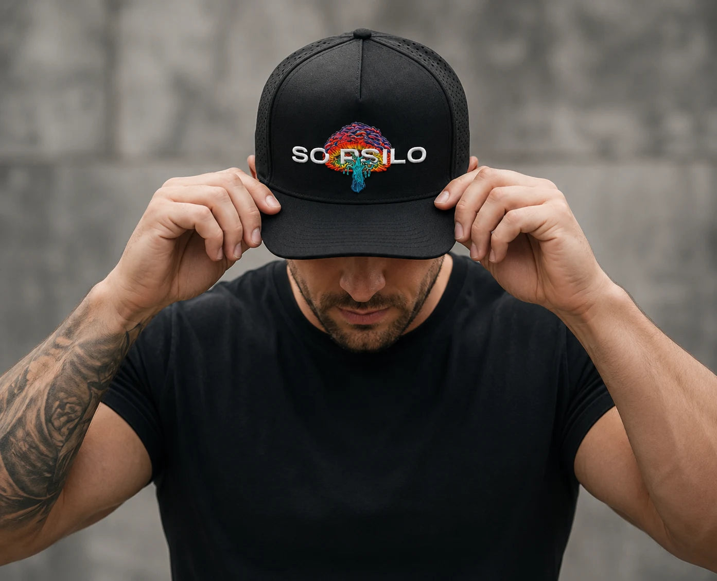 Close-up of a muscular man adjusting a black SOPSILO hat with vibrant mushroom embroidery against an outdoor urban background.